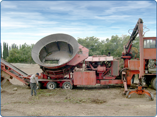 This machine is one of the major work horses of a green waste facility. This is used for wet materials. Wet materials would include cut tree branches, shrubs, and assorted vegetation.  The mechanical arm at the right side of this photo is used to pick up the materials (see previous image) and put them into the large round tub in the center of this picture. Then, the machine grinds up the materials to be further processed in the next stage.